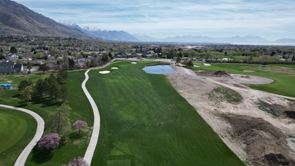 Drone view of a golf course fairway with pond, sand bunkers, paths, and snowcapped mountains.