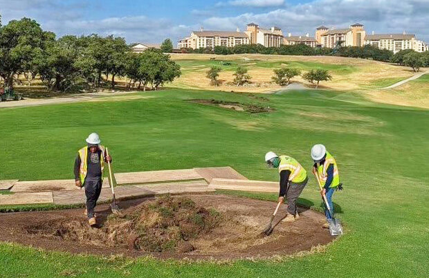 Three workers in safety vests repair a golf course bunker with a resort hotel in the background.