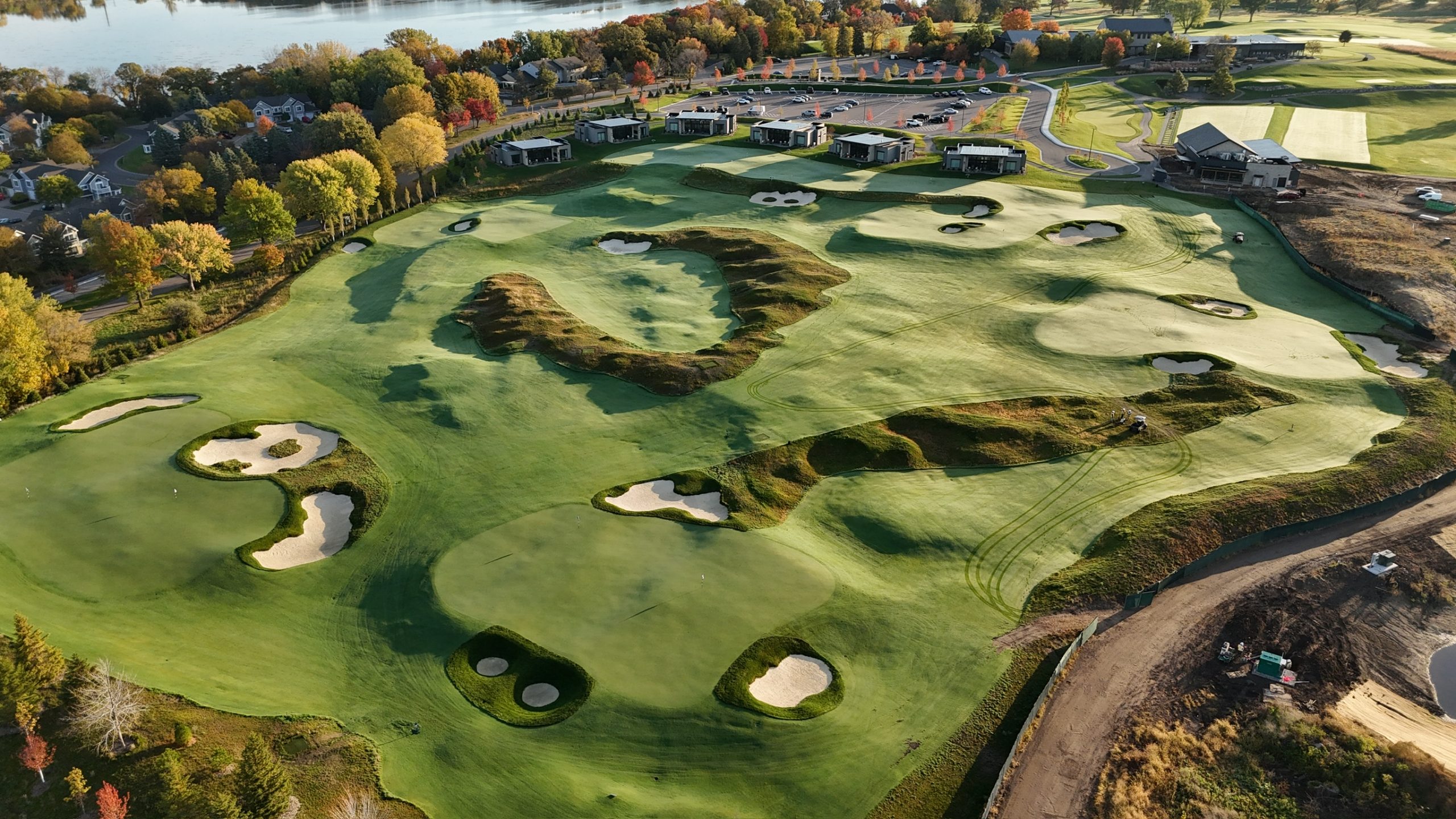 Aerial view of a lakefront golf course with rolling greens, sand bunkers, clubhouse, and autumn trees