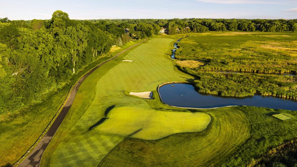 Drone view of lush golf course green with fairway, bunkers, and a pond beside wetlands