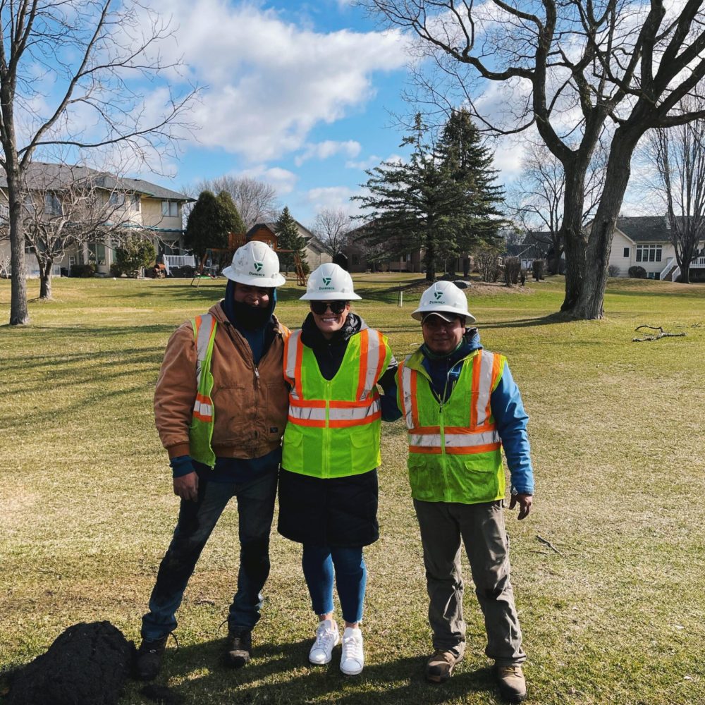 Three utility workers in safety vests and hard hats standing on a grassy residential lot